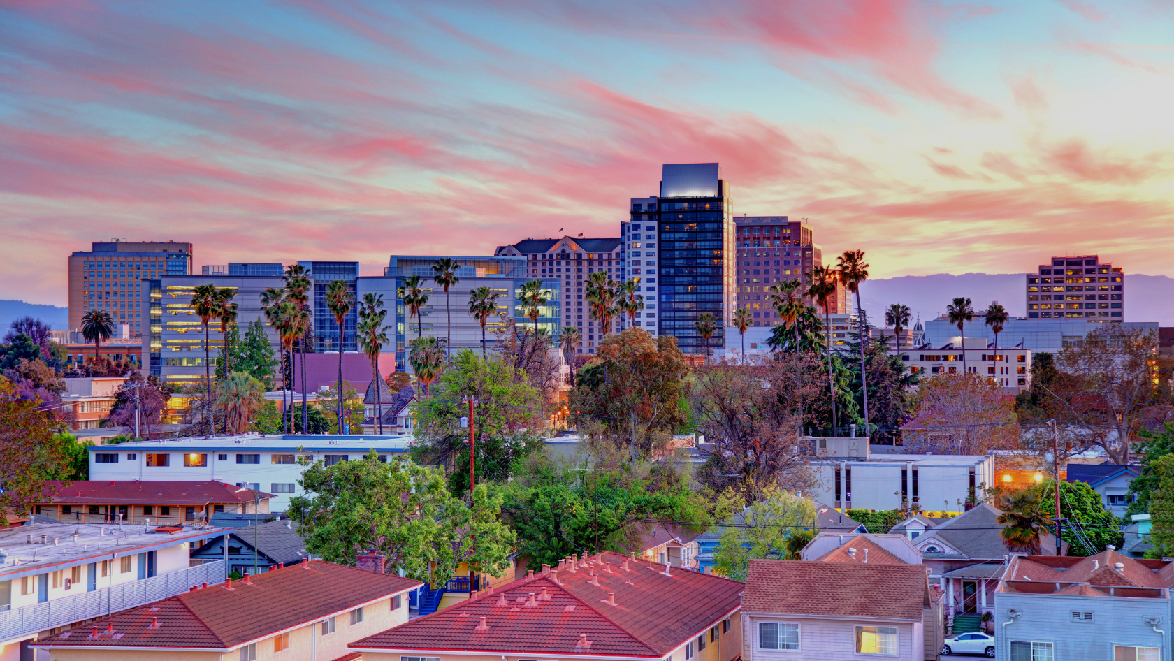 Sunset over a downtown skyline with tall buildings and palm trees beyond a residential neighborhood.