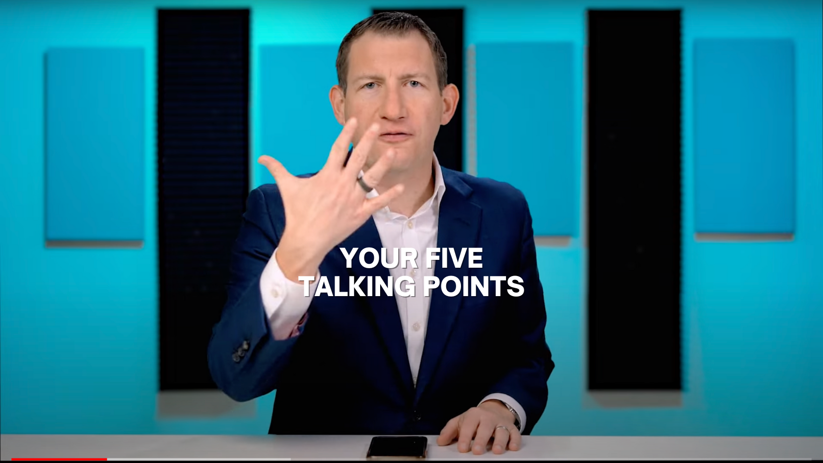 Presenter in a navy suit raises his hand while seated at a desk; bold text reads 'Your Five Talking Points' over a turquoise studio backdrop.