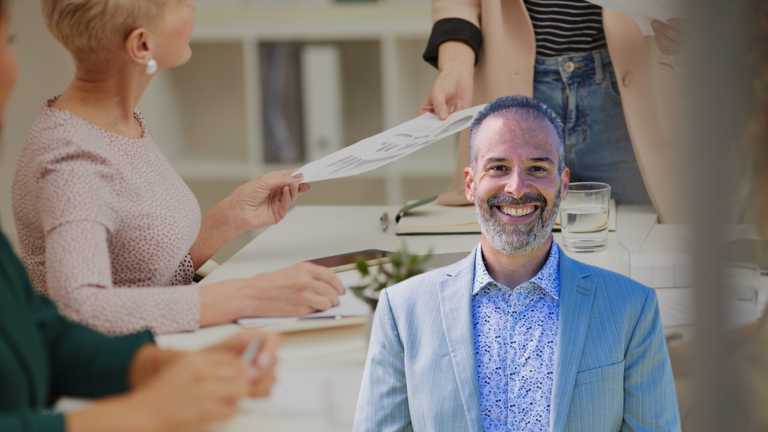 Smiling man in a light blue blazer sits at a desk as colleagues hand him a document across the table.