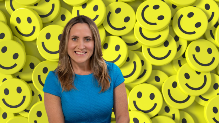 Woman in a blue shirt smiles in front of a wall of yellow smiley faces behind her.