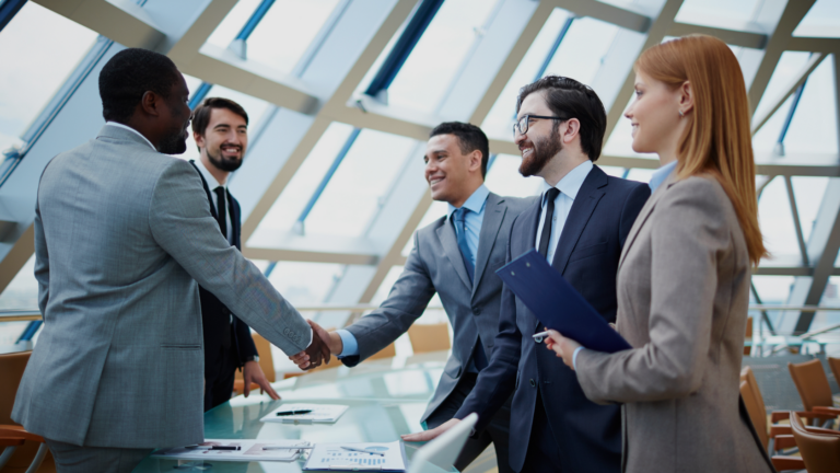 A diverse group of professionals in suits shake hands around a glass conference table in a modern office setting.