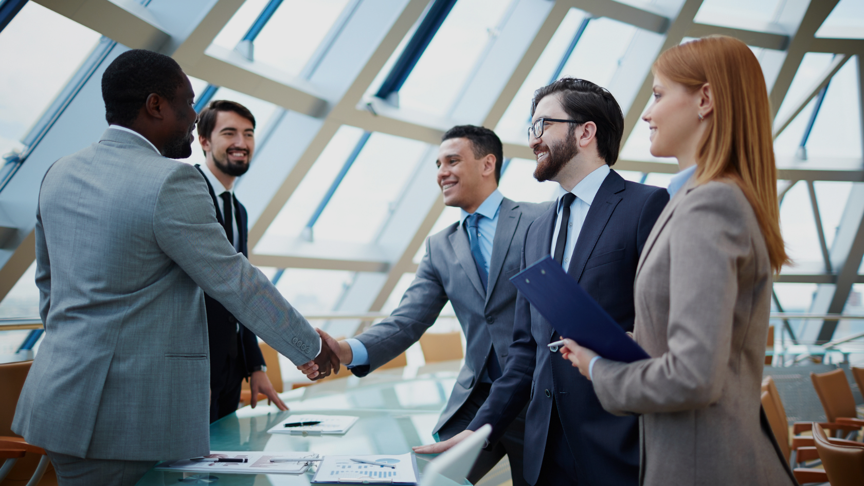 A diverse group of professionals in suits shake hands around a glass conference table in a modern office setting.