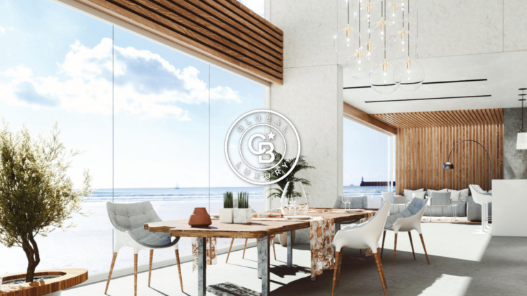 Elegant seaside lounge with large glass walls, wooden slat ceiling, a round rustic table and modern white chairs overlooking the ocean.
