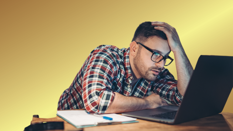 Man in a plaid shirt and glasses rests his head on his hand while looking at a laptop on a desk with a notebook nearby.