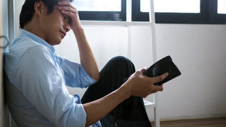 Man in a light blue shirt sits on the floor, head in hand, looking distressed while holding a black wallet.