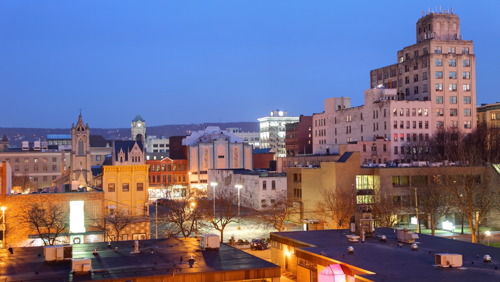Dusk city skyline with historic buildings, lit windows, and bare trees along the foreground rooftop line under a blue sky