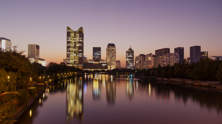 Sunset city skyline over a calm river with illuminated buildings reflecting on the water's surface.