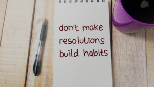 Notebook on a wooden desk shows the message: 'don't make resolutions build habits' with a pen to the left and a purple mug to the right.