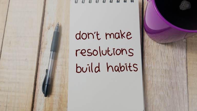 Notebook on a wooden desk shows the message: 'don't make resolutions build habits' with a pen to the left and a purple mug to the right.