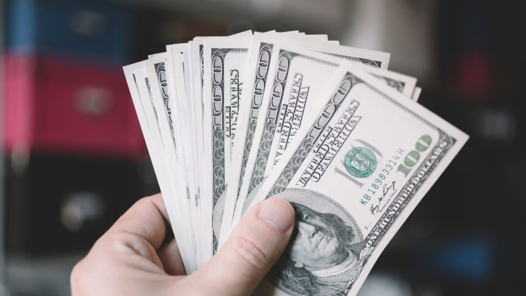 A hand holding a fanned stack of US $100 bills with the front note visible at the right edge.