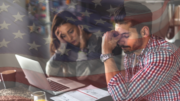 Man at a desk with a laptop, head in his hand, looking stressed; a woman blurred in the background with a faded American flag overlay.
