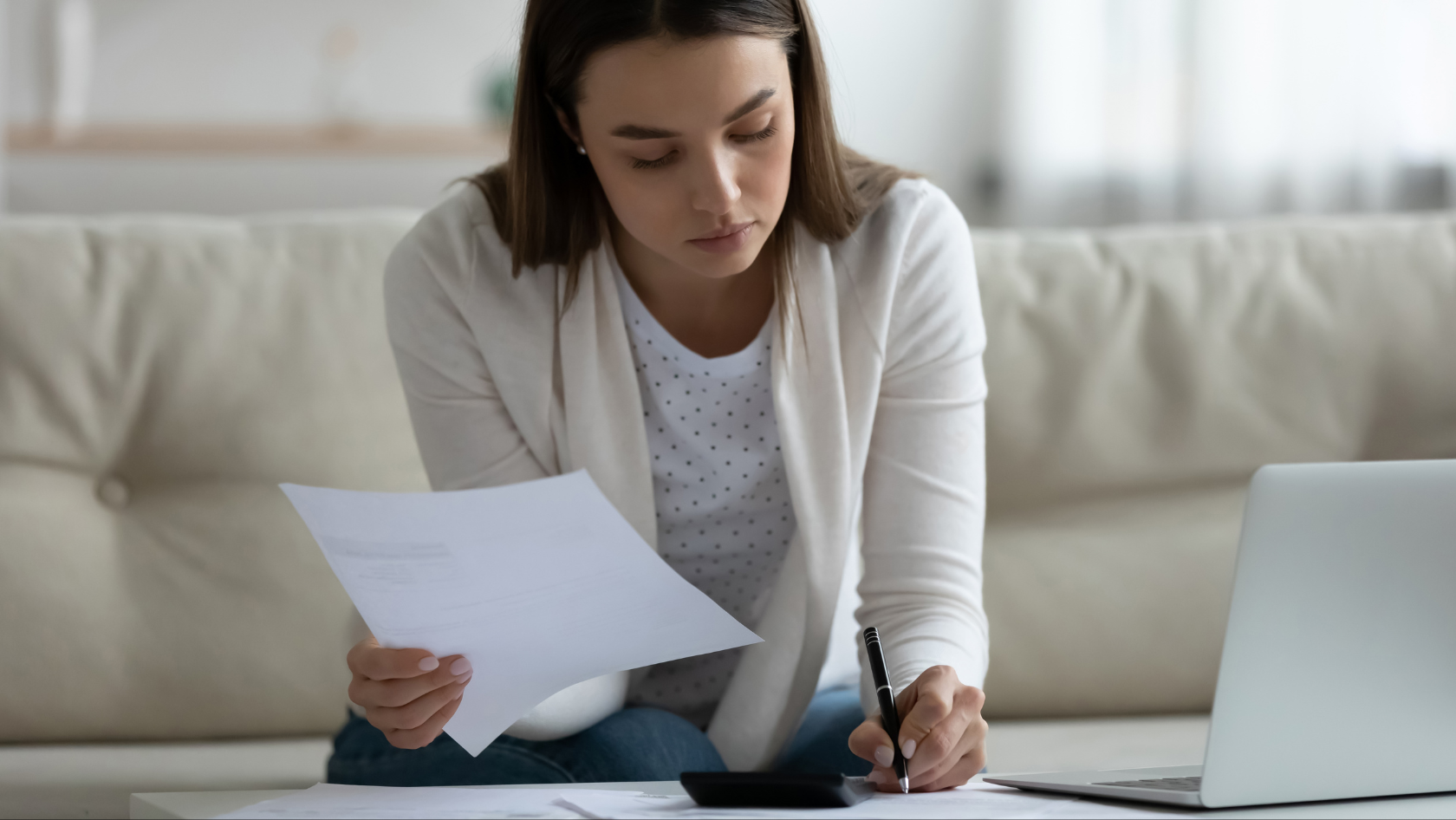 Woman sits on a sofa, reviewing papers with a pen while a laptop rests on a side table nearby.