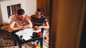 Two men sit on a rug at a low table, reviewing papers and notes while one shows something on his phone.