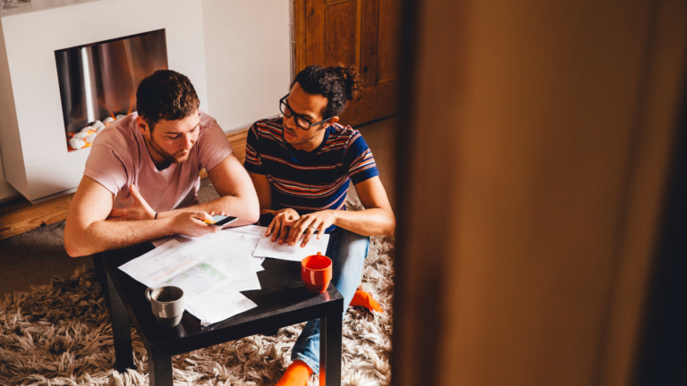 Two men sit on a rug at a low table, reviewing papers and notes while one shows something on his phone.
