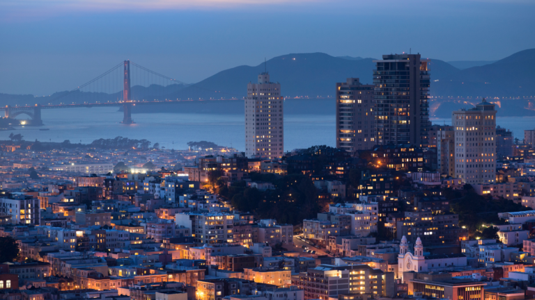 Dusk city skyline with the Bay Bridge illuminated across the bay and a dense grid of lit buildings in the foreground.
