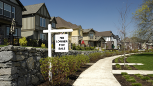 White 'No Longer For Sale' real estate sign on a post beside a stone wall in front of a row of suburban houses on a sunny street.