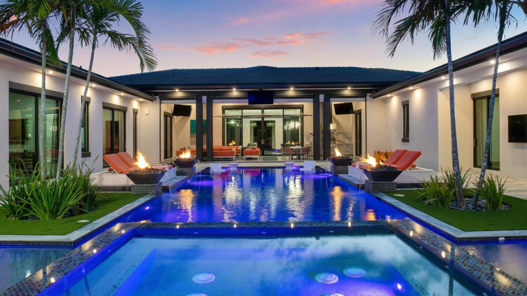 Luxurious courtyard pool area at dusk with blue-lit pool, palm trees, and orange lounge chairs around fire bowls and seating areas.