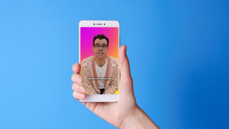 Hand holding a smartphone showing a man with glasses on a pink-to-orange gradient screen, with caption 'We have a lot of exciting things'.