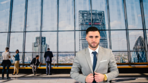 Man in a gray suit and navy tie stands in the foreground of a glass-walled office lobby with the city skyline behind him.