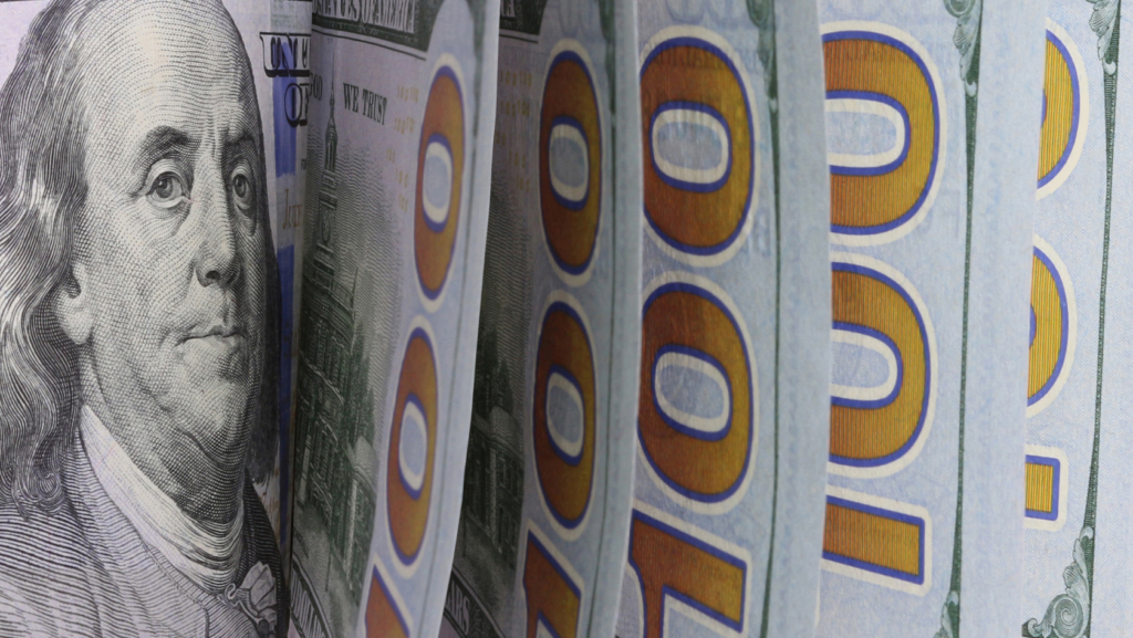 Close-up of fanned US hundred-dollar bills, with Benjamin Franklin on the left and large blue '100' numerals visible across the stack.