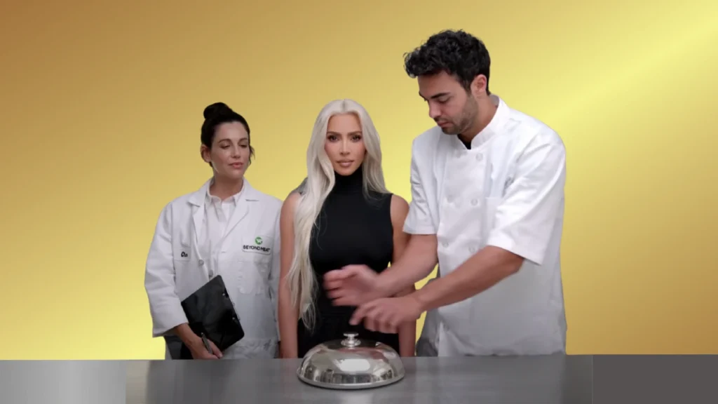 Three presenters stand around a silver cloche on a counter, with a yellow studio backdrop and two in white lab coats flanking a woman in a black top.