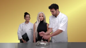 Three presenters stand around a silver cloche on a counter, with a yellow studio backdrop and two in white lab coats flanking a woman in a black top.