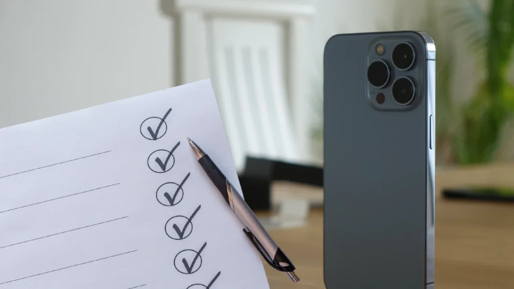 Checklist with checked boxes and a pen resting on a desk beside a modern smartphone on a wooden surface.
