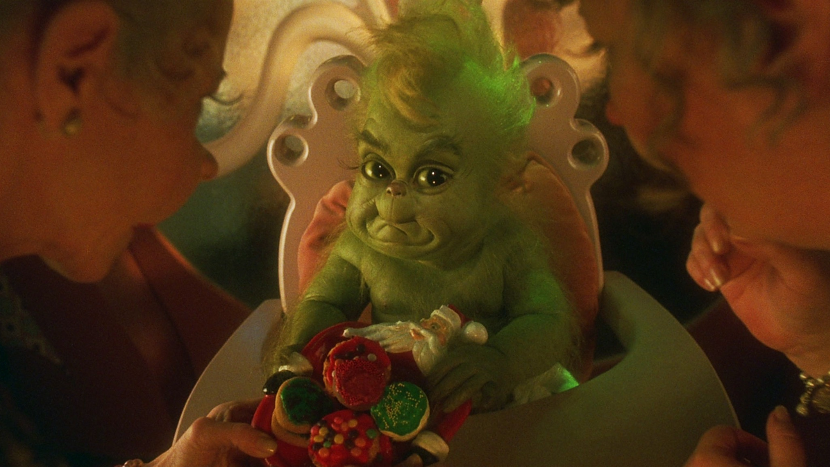 Green, furry baby creature sits in a high chair while adults present a colorful candy bouquet.