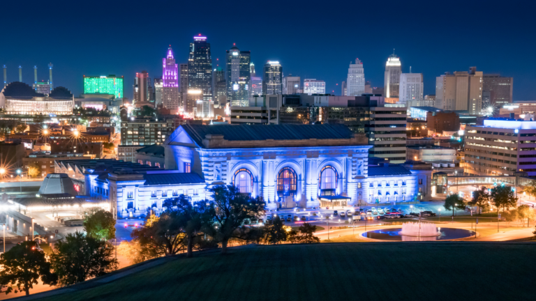 Nighttime cityscape with a blue-lit historic station in the foreground and a bright skyline behind.