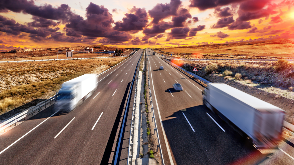 Highway with two lanes each side and a central barrier; trucks and cars travel fast through a desert at sunset with dramatic clouds in the sky.