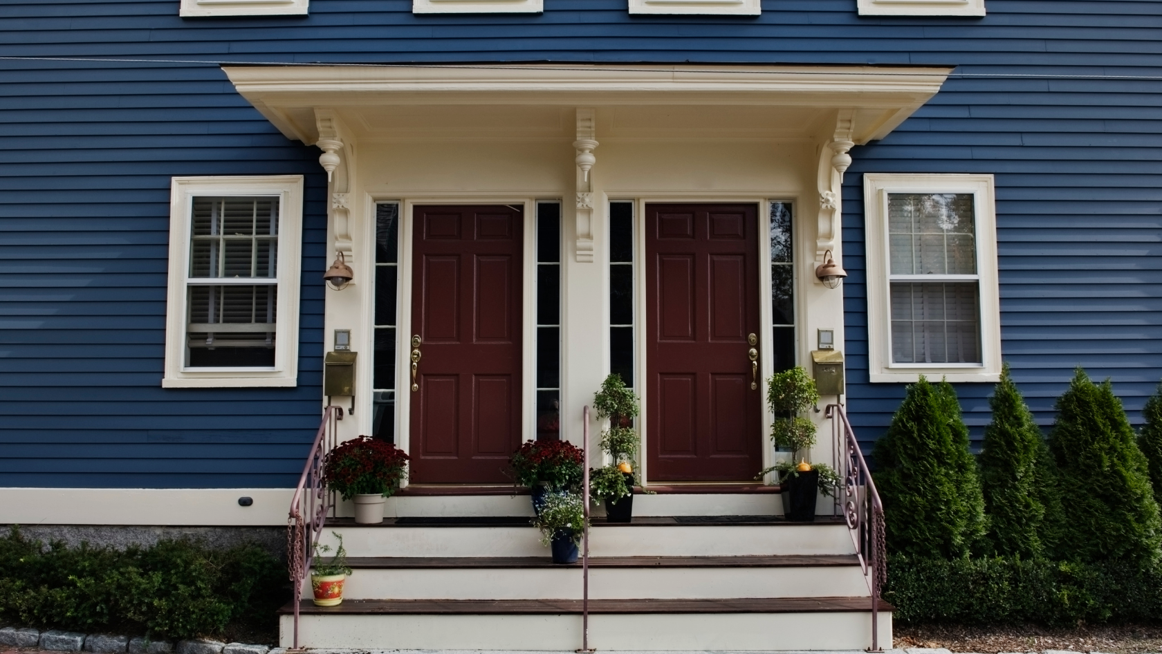 Blue house with white trim featuring two burgundy front doors, a small covered porch, and decorative columns, with potted plants on the steps.