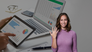 Smiling woman in a purple sweater sits beside a desk with a tablet and laptops displaying financial charts and graphs.