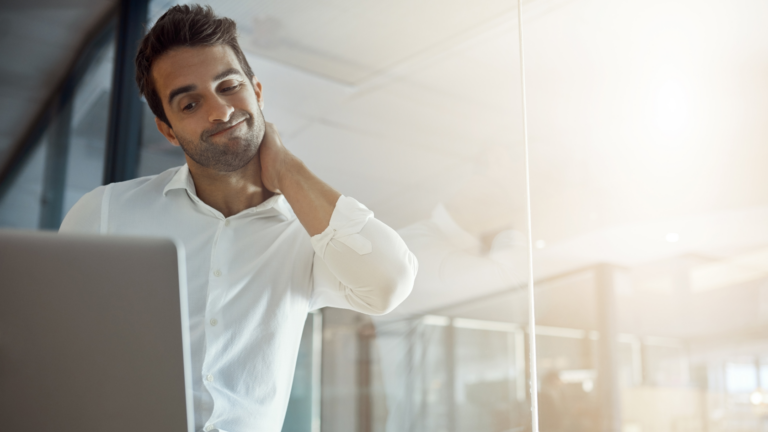Man in a white shirt stands by a glass wall, rubbing his neck while looking at an open laptop in a bright office.