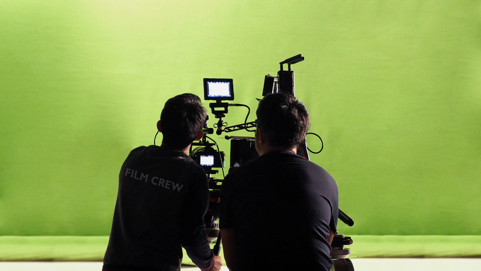 Two film crew members operate a camera rig in front of a bright green chroma key backdrop with a small monitor visible nearby