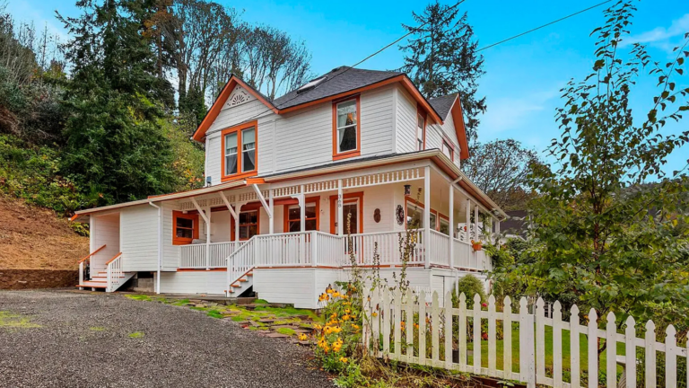 Two-story white house with orange trim and a wraparound porch, white picket fence, on a hillside with trees and a gravel driveway.
