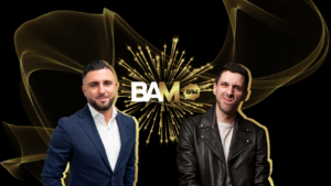 Two men in suits stand on either side of a glowing gold BAM logo with a starburst of fireworks on a black background.