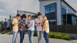 Five young adults standing outside modern townhouses, looking at their smartphones on a sunny day.