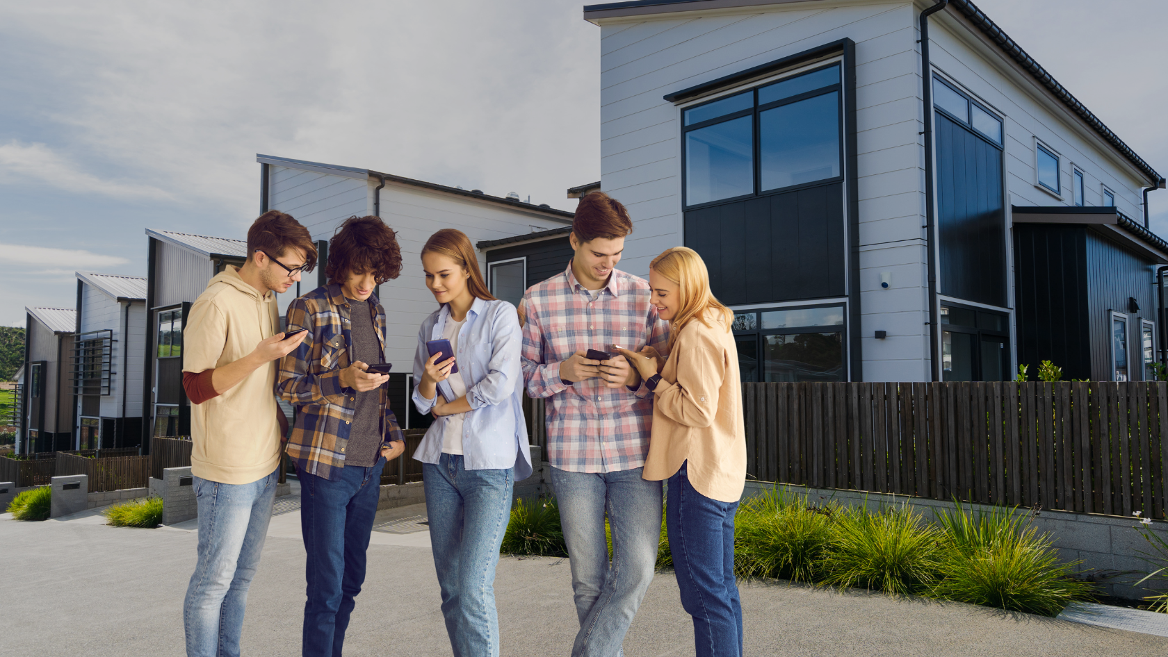 Five young adults standing outside modern townhouses, looking at their smartphones on a sunny day.