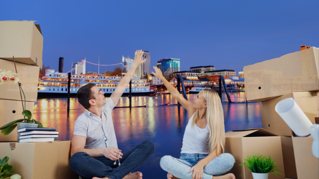 Couple sitting among packed cardboard boxes by a riverfront city, high-fiving in celebration of a move.