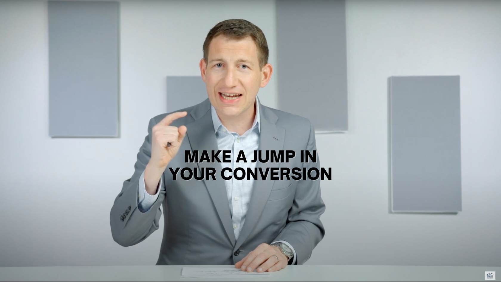 Business presenter in a gray suit sits at a desk, gesturing while a bold caption reads “MAKE A JUMP IN YOUR CONVERSION.”