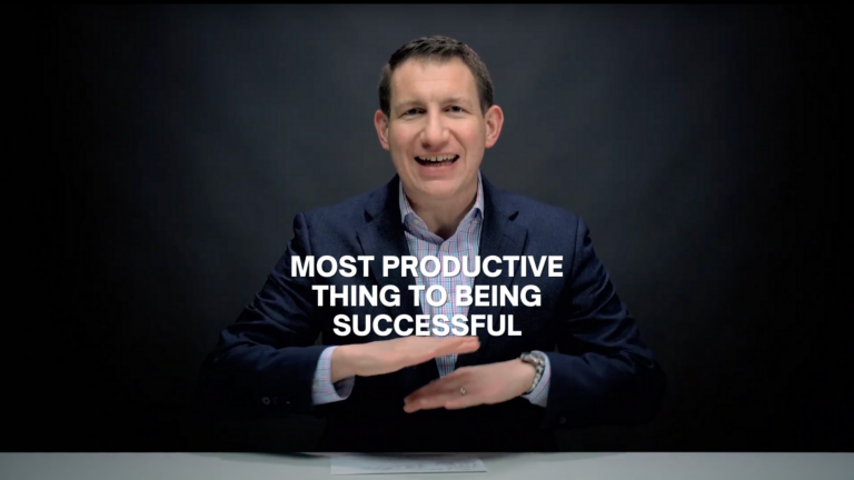Man in a dark blazer speaks to the camera at a desk; bold white text on screen reads “MOST PRODUCTIVE THING TO BE BEING SUCCESSFUL.”