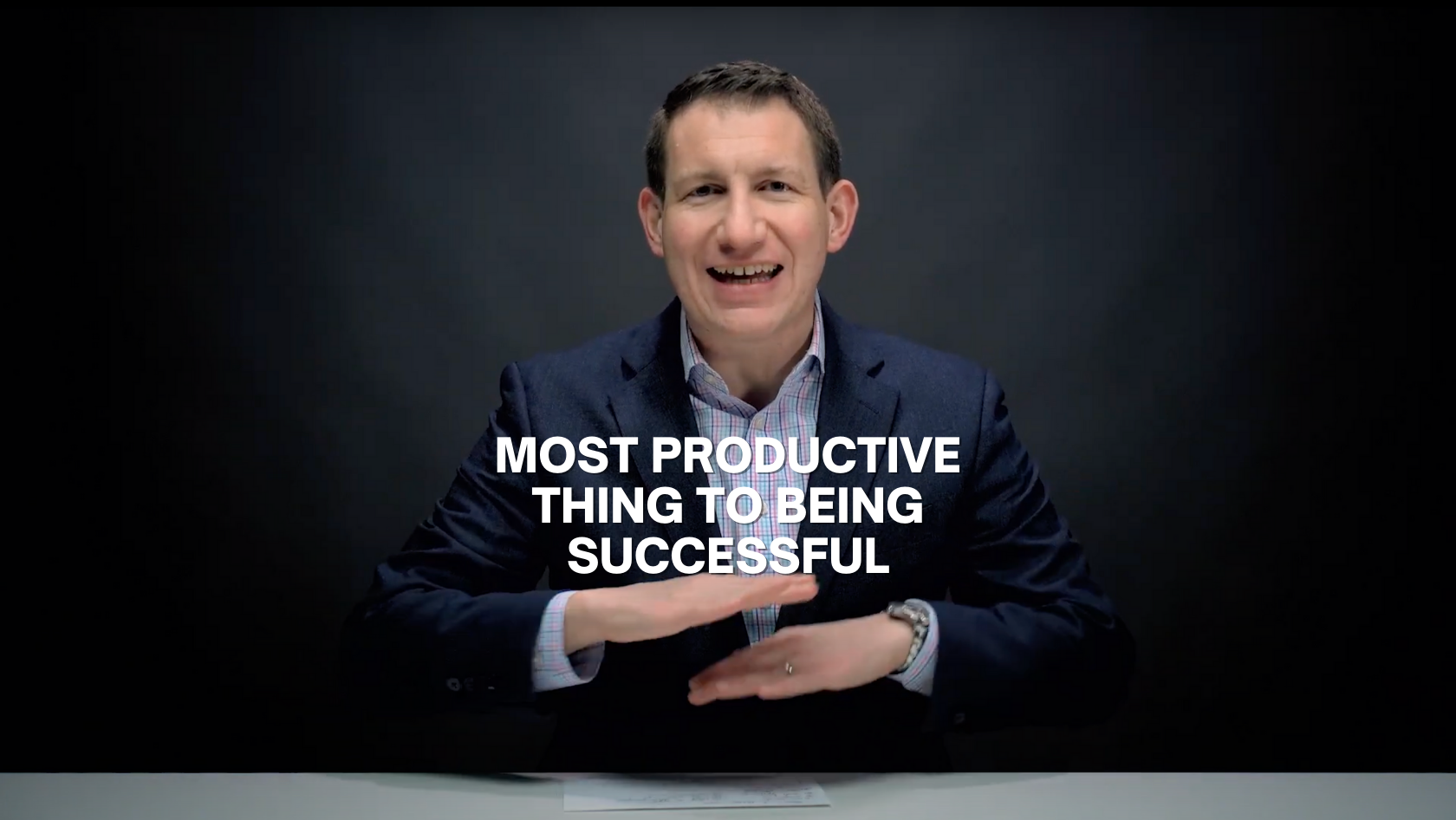 Man in a dark blazer speaks to the camera at a desk; bold white text on screen reads “MOST PRODUCTIVE THING TO BE BEING SUCCESSFUL.”