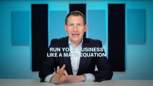 Professional man in a suit sits at a desk and speaks to camera; on-screen caption reads 'Run your business like a math equation'.