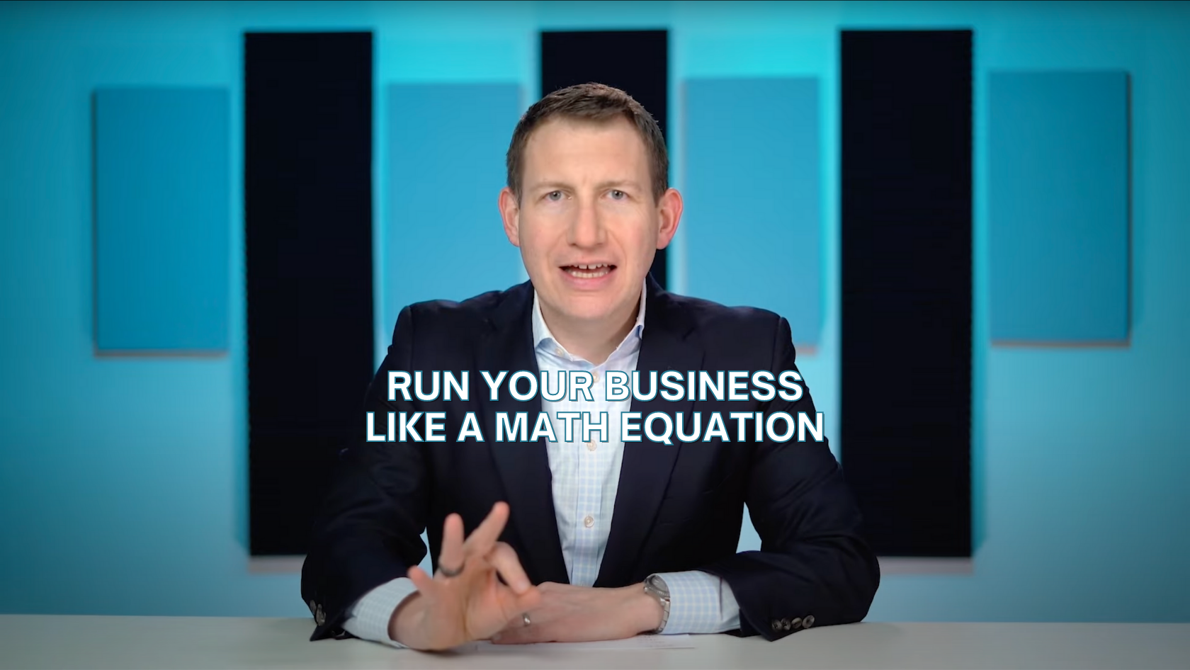 Professional man in a suit sits at a desk and speaks to camera; on-screen caption reads 'Run your business like a math equation'.
