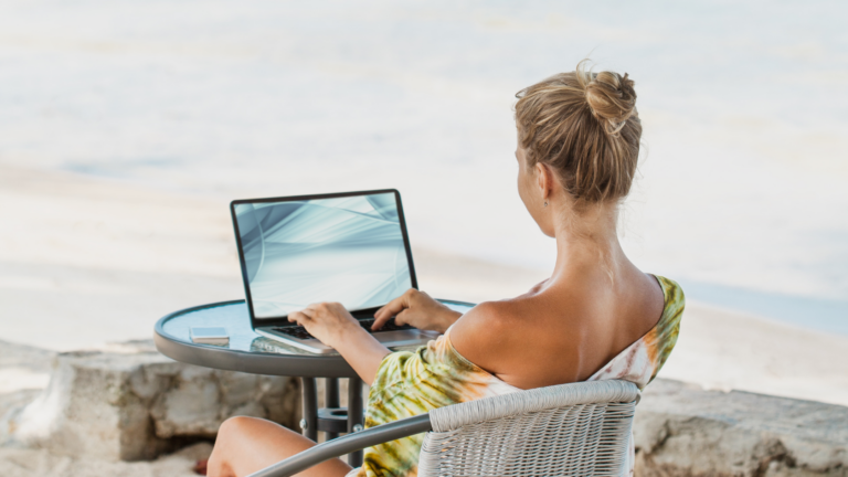 Woman sitting at an outdoor table by the water, typing on a laptop by the shore.