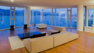 Modern living room with a beige sectional sofa, a glossy black coffee table, and a white ottoman, set on a light rug in front of expansive windows overlooking a harbor and distant mountains at dusk.