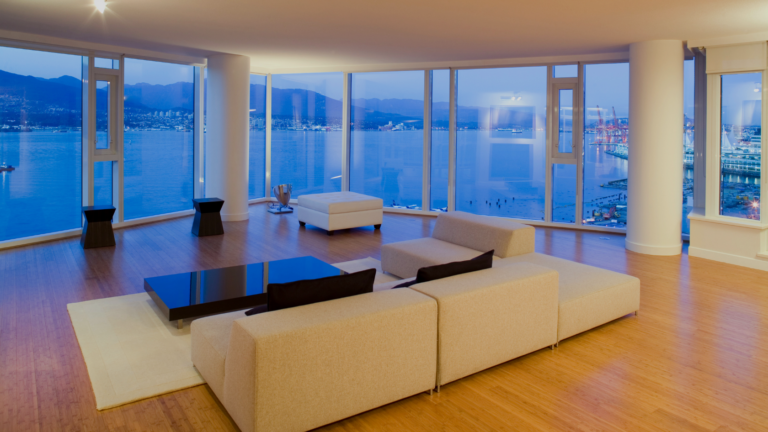Modern living room with a beige sectional sofa, a glossy black coffee table, and a white ottoman, set on a light rug in front of expansive windows overlooking a harbor and distant mountains at dusk.