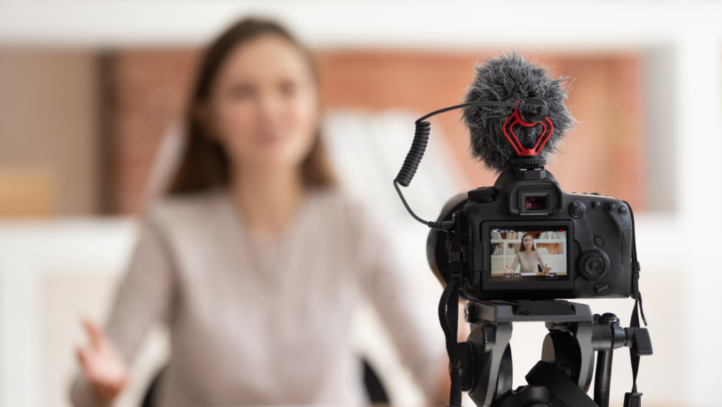 Blurred woman being interviewed in a studio while a DSLR on a tripod with a fluffy windscreen microphone records in the foreground, both facing her.