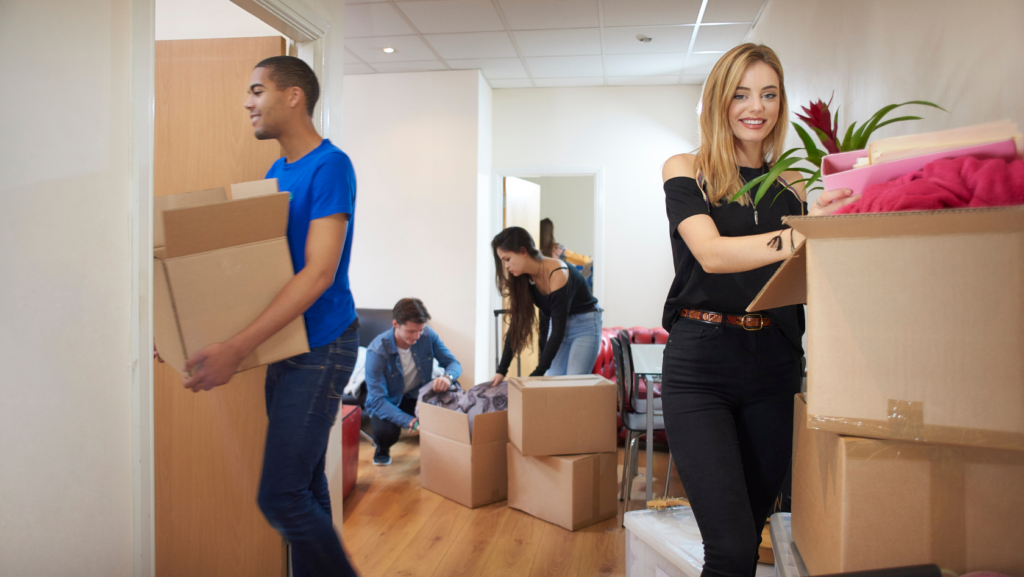 Smiling woman in a black top stacks items into a large box of pink blankets in a bright room with other volunteers and boxes nearby.