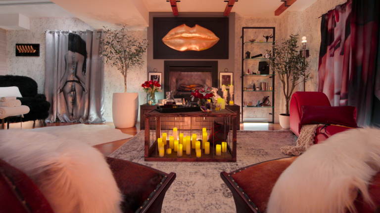 Lux lounge with candles on a central table and a copper lips sculpture above the fireplace, flanked by plants and shelves.